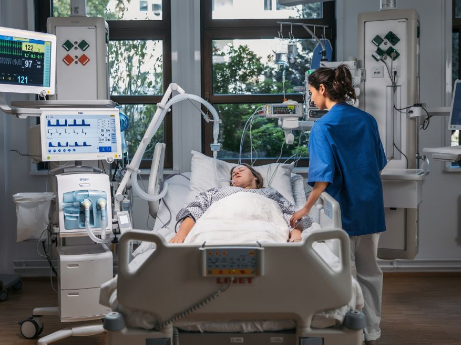 Pacient na lůžku intenzivní péče s monitorovací technikou a zdravotníkem.