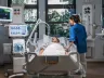 Pacient na lůžku intenzivní péče s monitorovací technikou a zdravotníkem.
